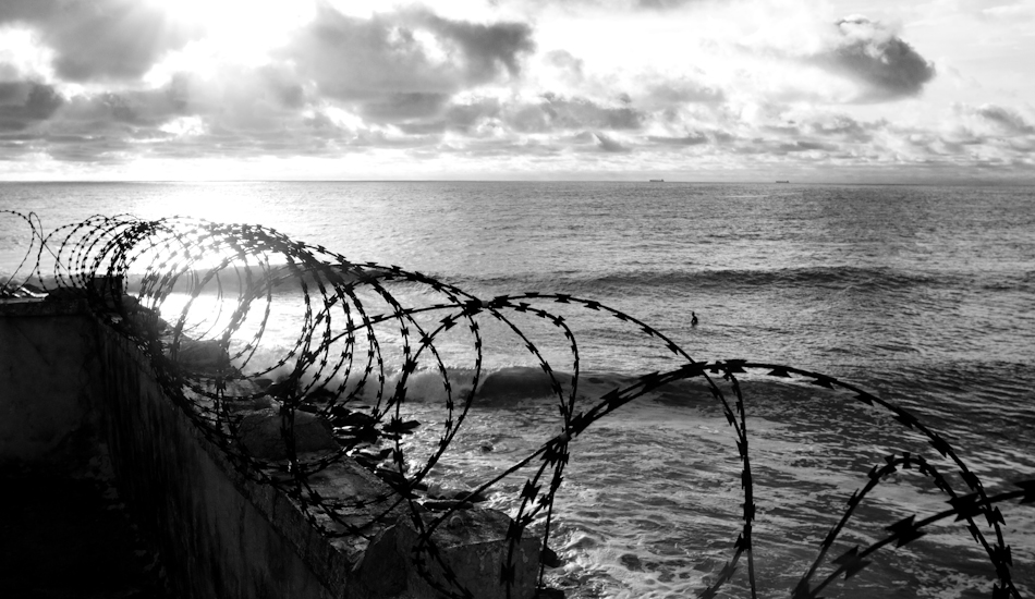 To surf this wave you have to bribe a security guard to let you through a metal trap door, aptly nick named “the poop shoot.” Then you cross a rocky jetty littered with human feces. Alfred Lomax, Liberia’s first surfer, sits alone out the back at Poopie Point. Photo: Brody/<a href=\"https://www.surfresource.org/\" target=_blank>SurfResource.org</a>
